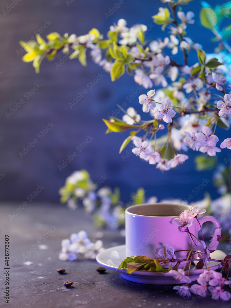 A white cup of coffee on the table with a branch of blooming white cherry. Spring greeting card . Neon colored light