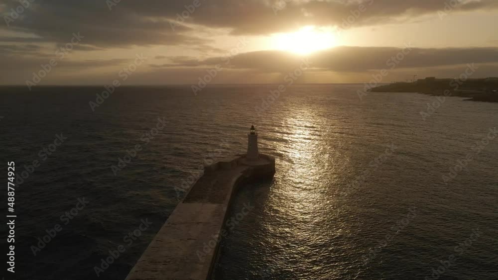 Cinematic aerial view over the light house during a scenic sunset with the sun light reflecting in the sea at St. Elmo Breakwater in Valletta, Malta (Drone footage) Cinematic aerial view over the light house during a scenic sunset with the sun light reflecting in the sea at St. Elmo Breakwater in Valletta, Malta (Drone footage)