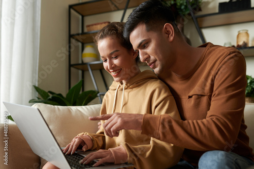 Young man pointing at the screen of laptop while sitting on sofa together with woman in the room