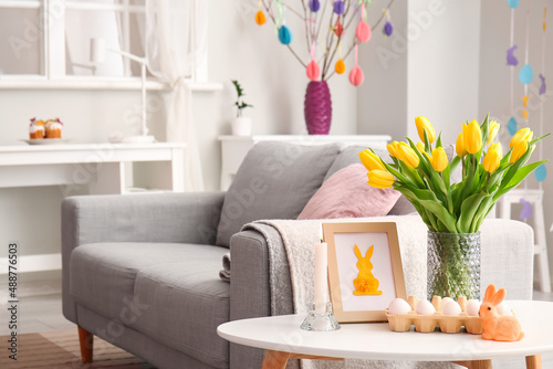 Holder with Easter eggs, photo frame, vase with tulips and burning candle near sofa in living room
