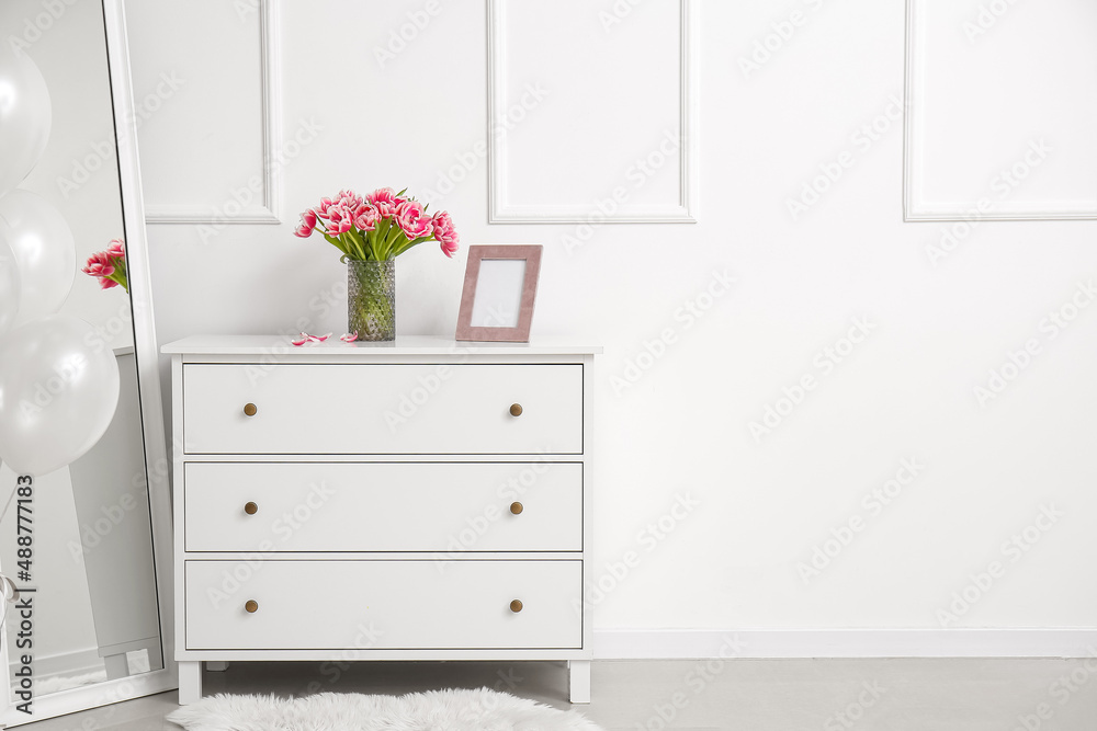 Vase with tulips and blank photo frame on chest of drawers near white wall in room Vase with tulips and blank photo frame on chest of drawers near white wall in room