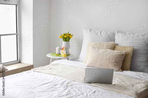 Comfortable bed with laptop, vase with tulips and Easter decor on table near light wall