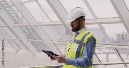 Close up of male chief engineer in hardhat and vest standing inside commercial building construction site using tablet to look at architectural plan of panoramic glass roof.