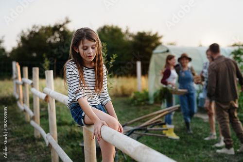 Fototapeta Naklejka Na Ścianę i Meble -  Little girl sitting on fence outdoors with her family at background at community farm.