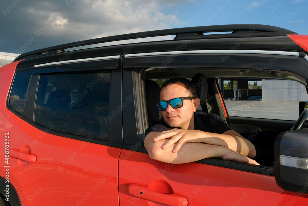 A young man in sunglasses sits in a car in a parking lot on a sunny day