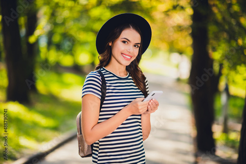 Photo of cute charming young woman dressed striped outfit cap backpack typing modern device walking smiling outside landscape
