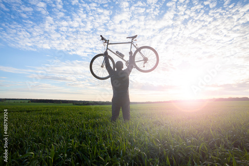 A cyclist holds a bike raised up in his hands in a field at sunset.