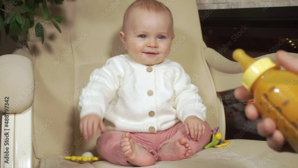 baby sits on armchair and starts to smile when hand of father holds out bottle