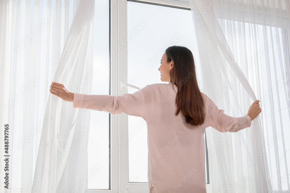Morning of pretty young woman opening curtains in room