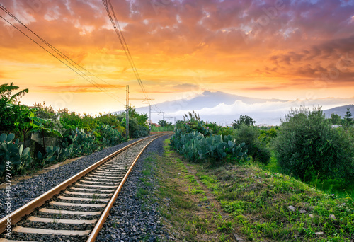 old evening railroad leading to a sunset glow in mountains with green bushes and cjlorful cloudy sku on the background