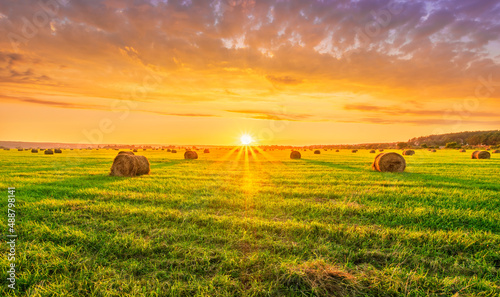 Scenic view at picturesque burning sunset in a green shiny field with hay stacks  bright cloudy sky   golden sun rays and road leading far away  summer valley landscape
