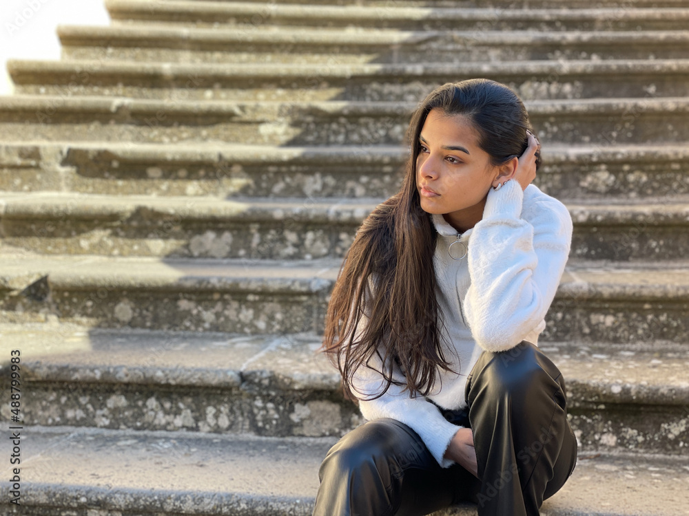 Sad looking young woman sitting outside and holding her head while looking away Sad looking young woman sitting outside and holding her head while looking away