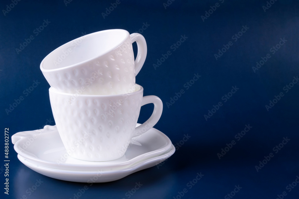 Two beautiful white cups (mugs) in the shape of strawberries and two saucers are stacked on top of each other on a dark blue background. Empty clean dishes. Space for text. Two beautiful white cups (mugs) in the shape of strawberries and two saucers are stacked on top of each other on a dark blue background. Empty clean dishes. Space for text.