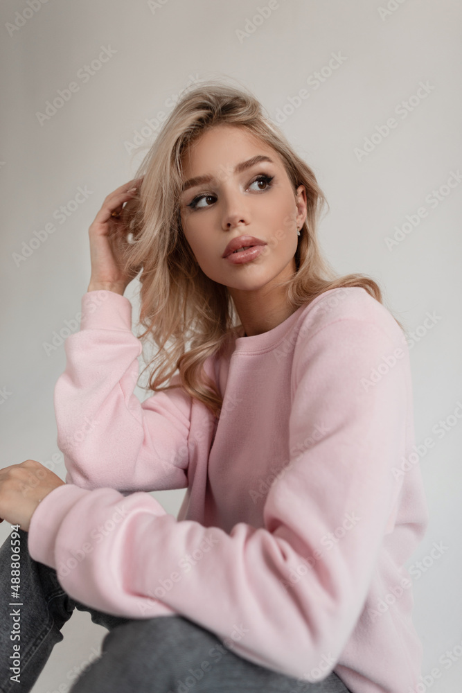 Glamorous young stylish girl with blonde hair in a pink fashionable sweatshirt and vintage jeans sits in the studio on a white background