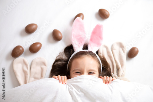 Happy Easter. cute beautiful girl with blue eyes and bunny ears peeking out from under the blanket. Chocolate eggs are scattered on the bed. Egg hunt. Lifestyle