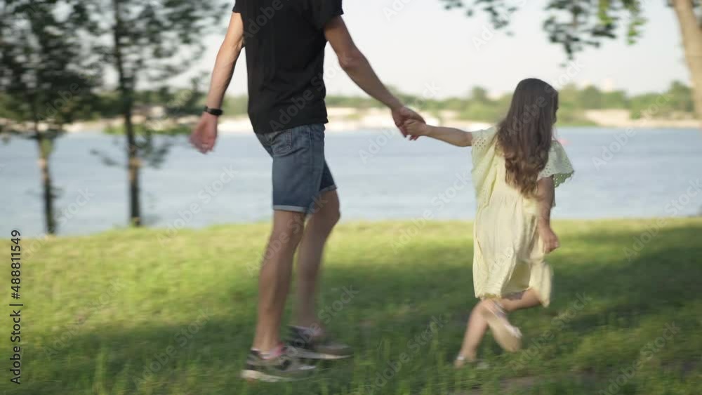 Wide shot joyful girl running in sunny park holding hand of young man. Panning shot of cheerful Caucasian daughter having fun enjoying weekend leisure with father outdoors. Childhood and family Wide shot joyful girl running in sunny park holding hand of young man. Panning shot of cheerful Caucasian daughter having fun enjoying weekend leisure with father outdoors. Childhood and family