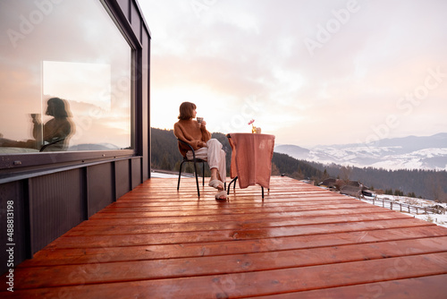 Woman having a breakfast while resting on terrace of tiny house in the mountains, enjoying beautiful landscape during sunrise. Concept of small modern cabins for rest and escape to nature