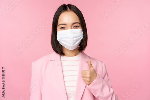 Asian businesswoman in suit and medical face mask, showing thumbs up, recommending wearing personal protective equipment in office during covid-19 pandemic, pink background photo