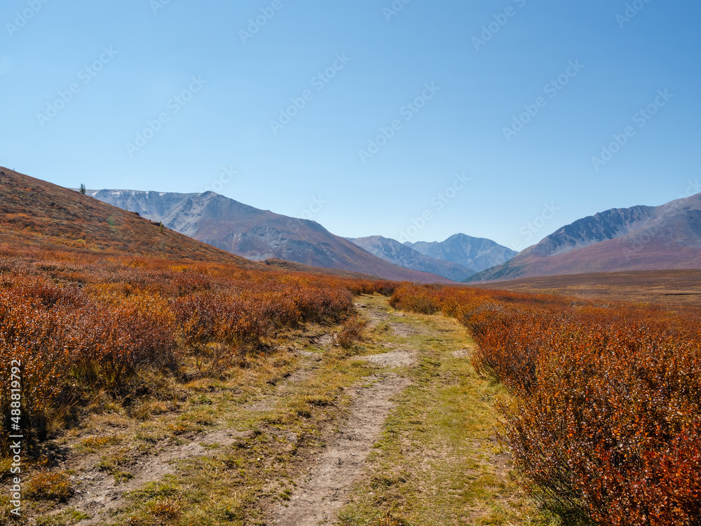 Dirt road through the autumn mountain plateau. Rough dirt road leading through orange autumn fields into mountains beyond under blue sky. Dirt road through the autumn mountain plateau. Rough dirt road leading through orange autumn fields into mountains beyond under blue sky.
