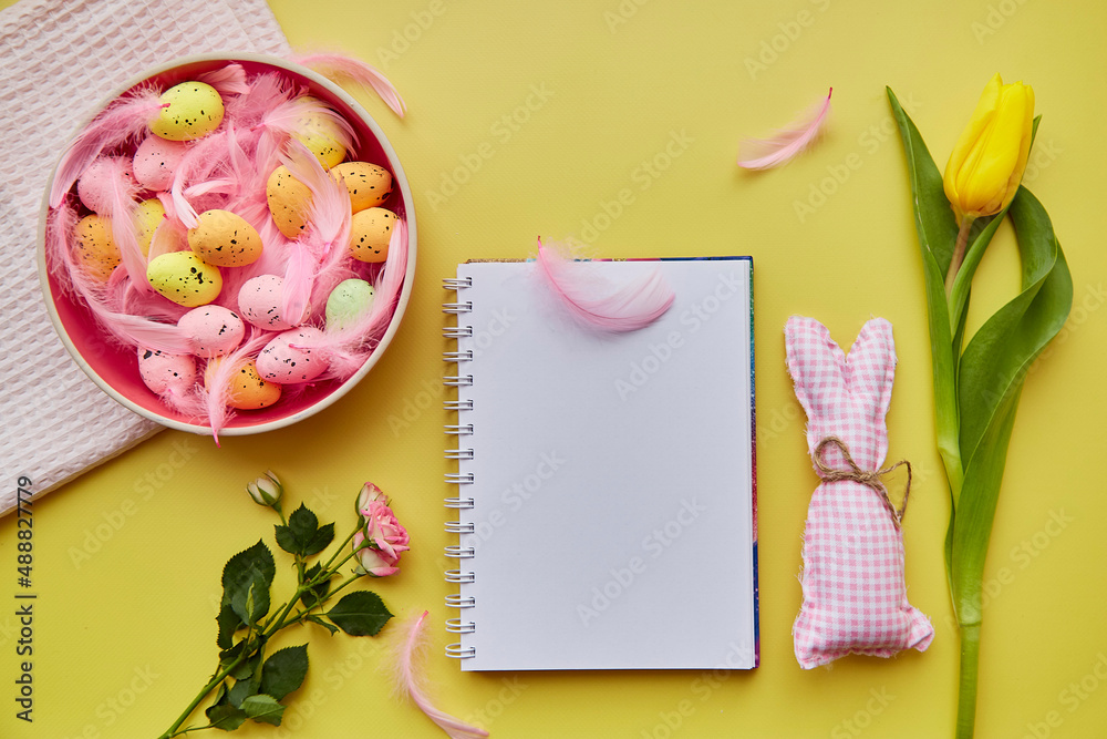 Easter mock up notepad. Pink bowl with colorful eggs, pink feathers, crafting cute bunny and flowers. Happy Easter concept. Top view. Post card mock up on yellow background