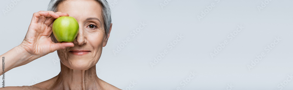 smiling middle aged woman covering eye with green apple isolated on grey, banner smiling middle aged woman covering eye with green apple isolated on grey, banner