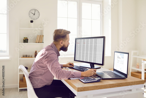 Man working in office. Serious busy financial accountant sitting at desk with calculator, laptop and desktop computers, looking at screens and working with electronic spreadsheets and invoice files photo