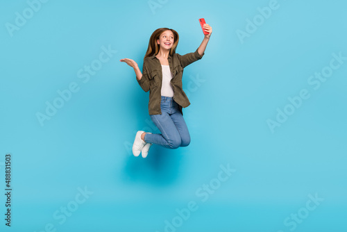 Full size photo of beautiful teen girl jump talk on camera blogger hold hand advertise isolated on blue color background