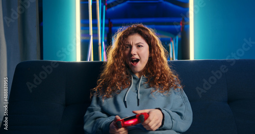 Excited funny young woman playing video games pressing buttons on wireless controller. Female gamer trying to win the competition. Room with neon lights on background.