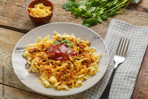 Pasta bolognese garnished with herbs and cheese in a plate on a wooden table next to a fork and a napkin and greens on a board and grated cheese.