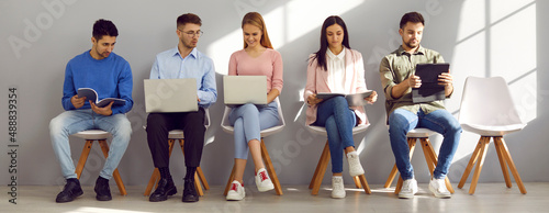 Young people holding paper documents, resumes and laptops are sitting in line for interview. Men and women in casual clothes communicate and prepare for business meeting at staffing agency. Banner.