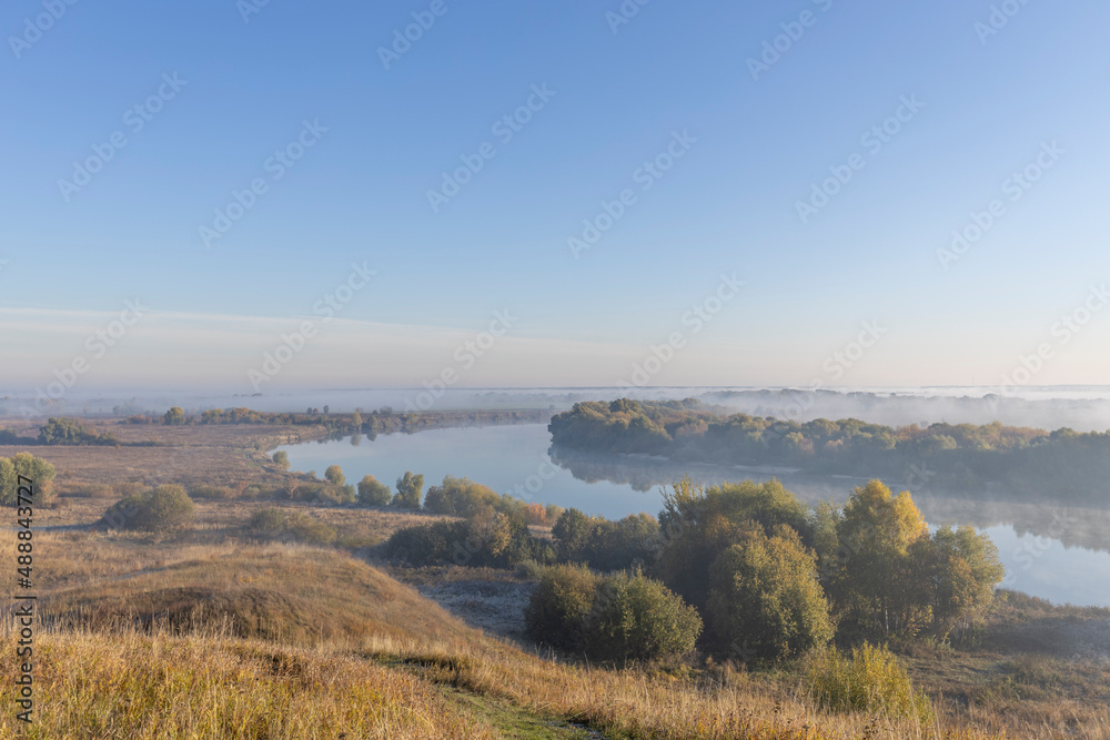 Autumn landscape in the early morning overlooking the river. A wide river and endless expanses of fields. Yellow leaves on trees and bushes are illuminated by the rays of the rising sun.
