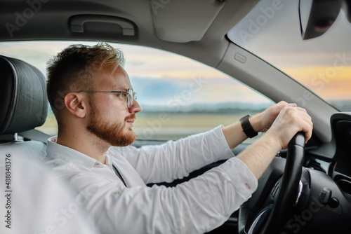 young male driver of a car in glasses and business clothes, rides at sunset. Business class driver concept