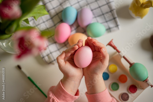 easter egg in the hands of a little girl over a table with painted eggs, watercolors and tulips. © InfiniteStudio