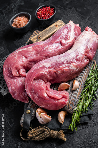 Pork tenderloin meat, raw uncooked fillet on butcher board with meat cleaver. Black background. Top view