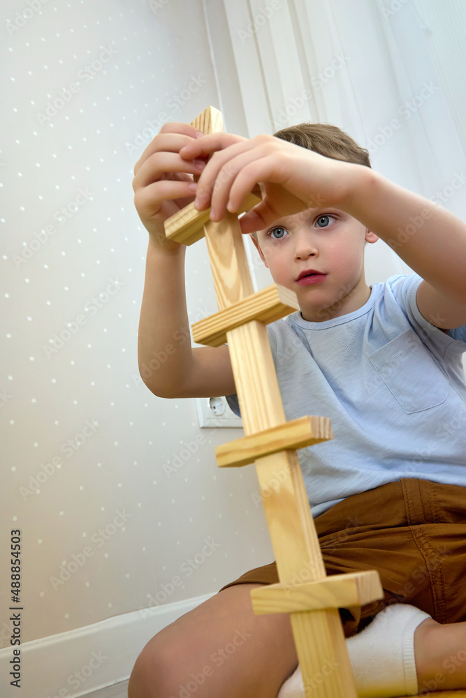 Obraz premium Cute Caucasian child diligently builds a tower of wooden bars. A boy in casual clothes plays with the details of a jenga. Educational children's games. Vertical image