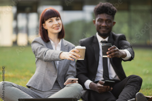 Close up serious businessman and businesswoman drinking take away coffee sitting on the grass outdoors. Portrait of business man and woman taking break with coffee to go outdoor.
