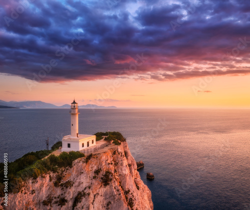 Lighthouse on the mountain peak at colorful sunset in summer. Aerial view. Beautiful lighthouse on the rock, sea and orange sky with purple clouds at dusk. Top view of Cape Lefkada, Greece. Landscape