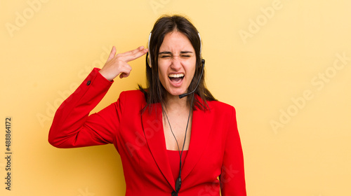 young hispanic woman looking unhappy and stressed, suicide gesture making gun sign. telemarketer concept