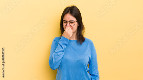 young hispanic woman feeling disgusted, holding nose to avoid smelling a foul and unpleasant stench