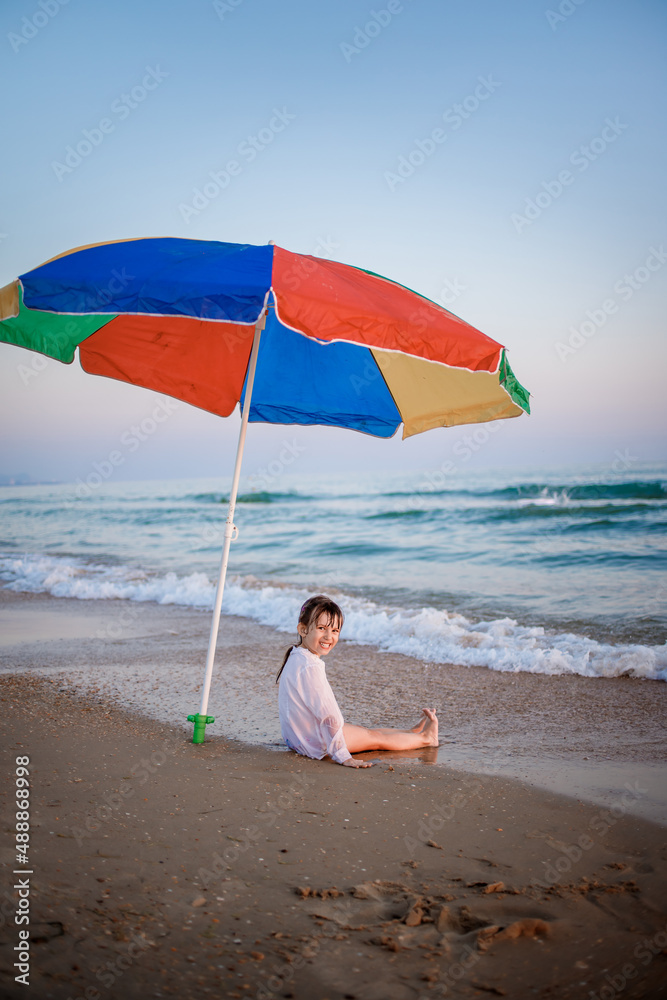 European child sits on the beach by the sea under a rainbow umbrella from the sun