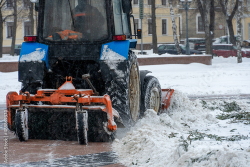 A tractor cleans snow in the city in winter after a snowfall. Cleaning city streets from snow. Tractor driver at work in the city square during a snowstorm.