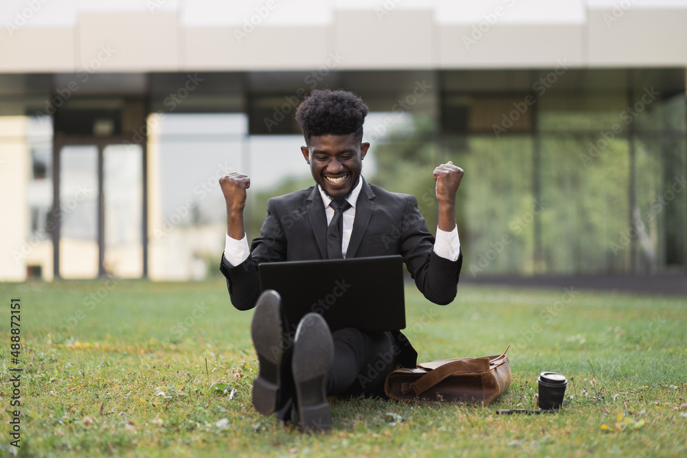 Office work and mindfullness. Handsome African young business man with raised hands working outside in front of company building, sitting on the grass with laptop and coffee cup. Well done, good news Office work and mindfullness. Handsome African young business man with raised hands working outside in front of company building, sitting on the grass with laptop and coffee cup. Well done, good news