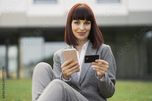 People, business, technologies, online payment. Beautiful smiling woman, sitting on grass outside, holds cellphone and credit card, makes shopping online or verifies account balance using mobile app