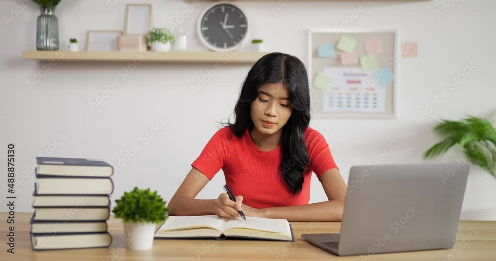 Portrait of Asian schoolgirl studying online making notes in copybook and waving hand via laptop at home. Distance learning and education concept. Portrait of Asian schoolgirl studying online making notes in copybook and waving hand via laptop at home. Distance learning and education concept.