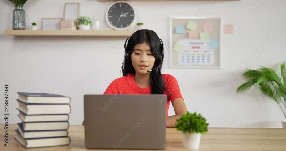 Portrait of Asian schoolgirl studying online making notes in copybook and waving hand via laptop at home. Distance learning and education concept. Portrait of Asian schoolgirl studying online making notes in copybook and waving hand via laptop at home. Distance learning and education concept.