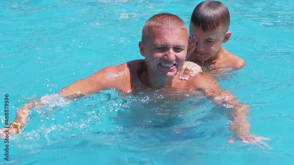 Happy Smiling Father with Son Swim in Open Transparent Swimming Pool. Man and a child are playing, splashing, having fun in the water on summer holidays. A family vacation at the resort, hotel.