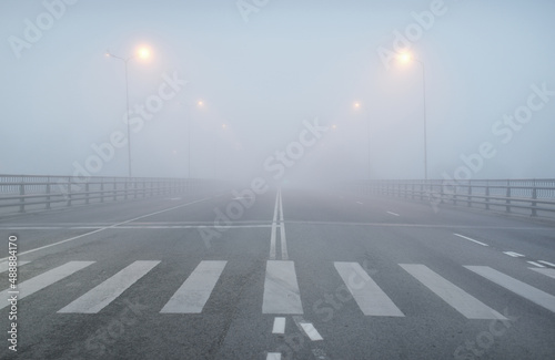 An empty illuminated asphalt road (highway) in a thick fog. Pedestrian walkway, crossing, street lights. Dangerous driving, walking, cycling, traffic laws concepts