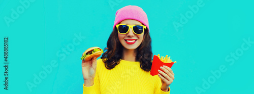 Portrait of stylish happy smiling young woman with burger and french fries fast food on blue colorful background
