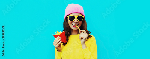 Portrait of stylish happy smiling young woman eating french fries fast food on blue colorful background
