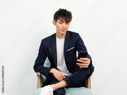 Portrait of handsome Chinese young man in dark blue leisure suit sitting in armchair and posing against white wall background. looking at mobile phone in hand with Legs crossed, young man lifestyle.